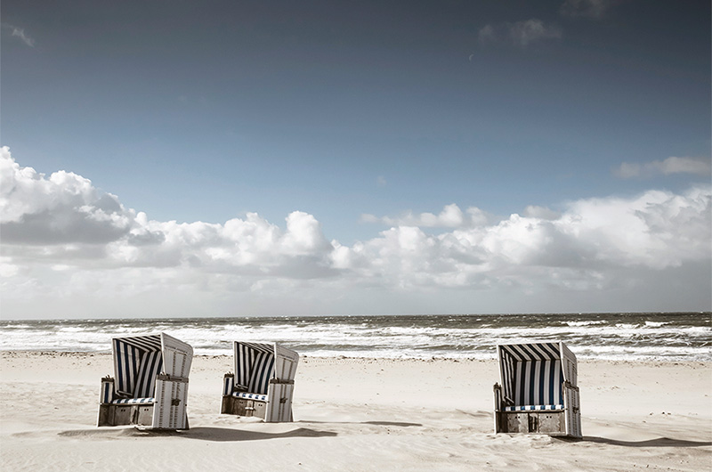 Sandstrand med tre blå- og hvit-stripete, doble strandstoler med vegger og tak.