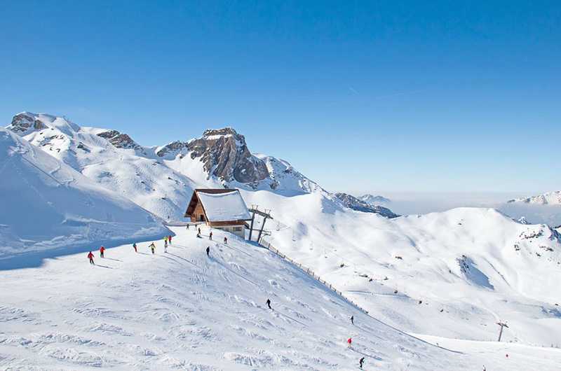 Skiheis med hus på toppen. Alpene i bakgrunnen, Folk på ski i forgrunnen.