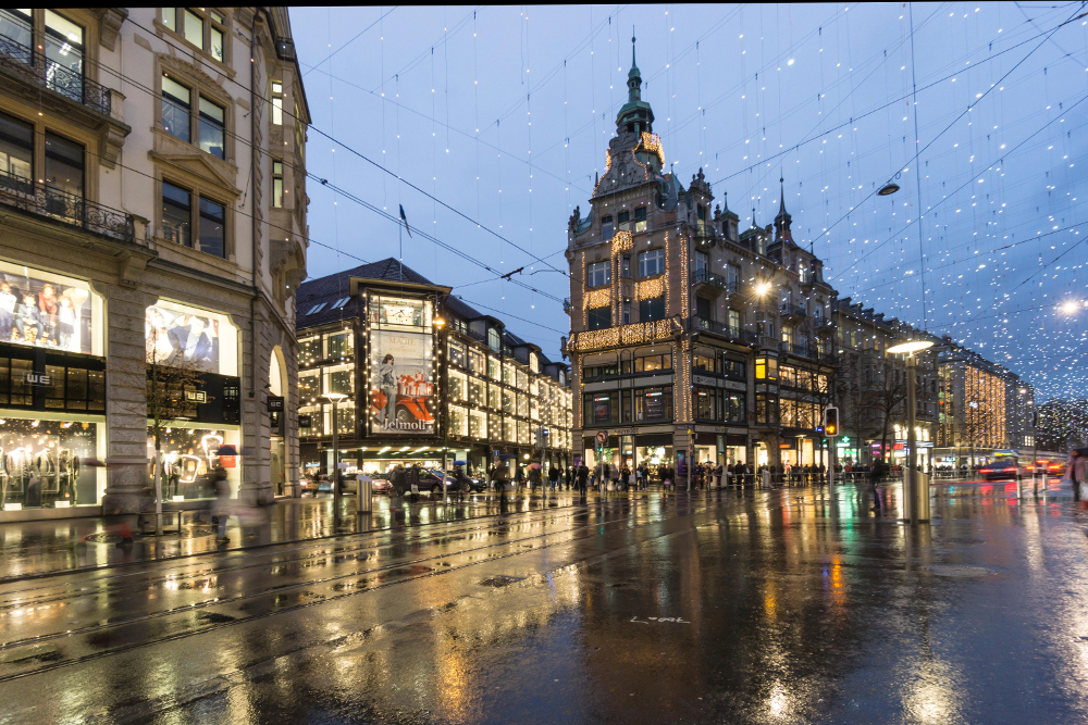 Regnvåt bygate med butikker og neonlys: Bahnhofstrasse i Zürich